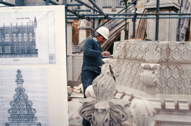 A worker wearing a hard hat is carving intricate designs on a large stone architectural piece. In the background, architectural blueprints and scaffolding can be seen, indicating a construction or restoration site.