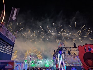A night scene featuring a vibrant display of fireworks in the sky with bright, colorful lights illuminating a stage and Ferris wheel. The atmosphere is lively with a clear view of a crowd enjoying the event, accompanied by festive decorations and a large cartoon character cutout in the foreground.