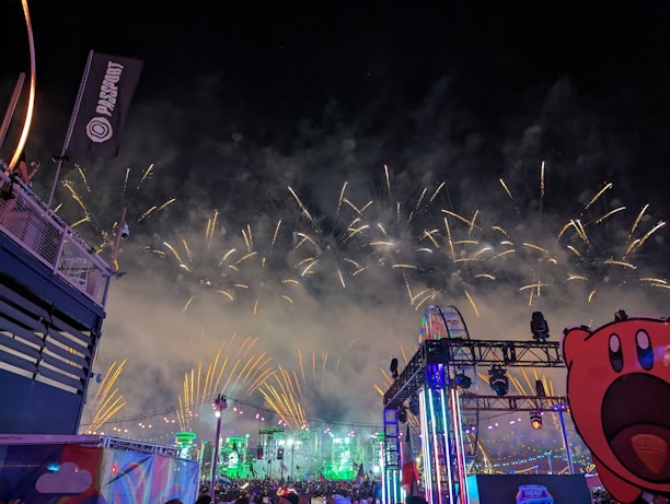 A night scene featuring a vibrant display of fireworks in the sky with bright, colorful lights illuminating a stage and Ferris wheel. The atmosphere is lively with a clear view of a crowd enjoying the event, accompanied by festive decorations and a large cartoon character cutout in the foreground.