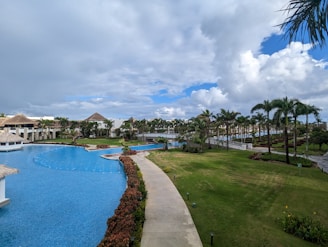 A luxurious resort setting featuring a large, winding swimming pool surrounded by manicured lawns and lush tropical palm trees. There are thatched-roof structures and white buildings in the background, with a partly cloudy sky overhead.