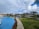 A luxurious resort setting featuring a large, winding swimming pool surrounded by manicured lawns and lush tropical palm trees. There are thatched-roof structures and white buildings in the background, with a partly cloudy sky overhead.