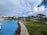 A luxurious resort setting featuring a large, winding swimming pool surrounded by manicured lawns and lush tropical palm trees. There are thatched-roof structures and white buildings in the background, with a partly cloudy sky overhead.