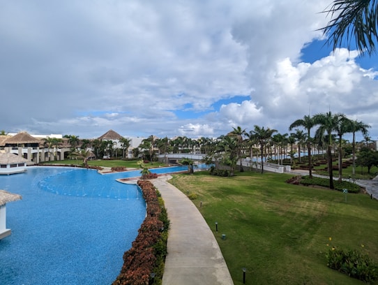 A luxurious resort setting featuring a large, winding swimming pool surrounded by manicured lawns and lush tropical palm trees. There are thatched-roof structures and white buildings in the background, with a partly cloudy sky overhead.