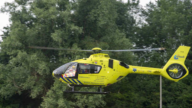 A bright yellow helicopter with ambulance markings hovers in front of a backdrop of dense green trees. The helicopter has blue accents and several logos and text on its side, indicating it is part of a mobile medical team.