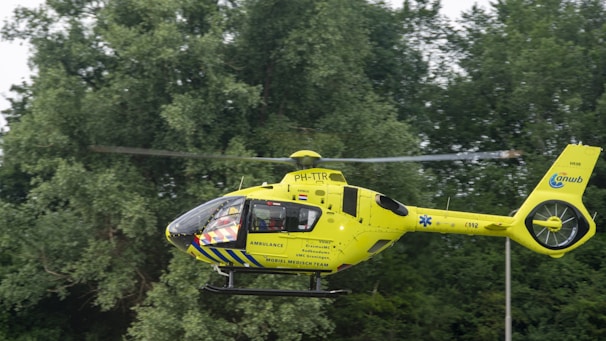 A bright yellow helicopter with ambulance markings hovers in front of a backdrop of dense green trees. The helicopter has blue accents and several logos and text on its side, indicating it is part of a mobile medical team.