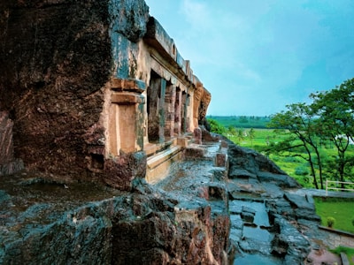 A historic stone structure is set amid a verdant landscape. The building features carved details and weathered stones, suggesting age and historical significance. The scene is complemented by lush greenery and a clear sky in the background, evoking a serene and timeless atmosphere.
