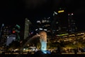 A night view of a cityscape featuring a prominent illuminated statue of a merlion with water flowing from its mouth. Skyscrapers with various brightly lit signs and logos form the backdrop under a dark sky. Green foliage appears in the foreground near the statue, adding contrast to the urban setting.