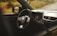 Interior view of a car showing the steering wheel with a prominent Suzuki logo, dashboard with air vents, gear shift, and side mirror. Outside, there is a grassy landscape.