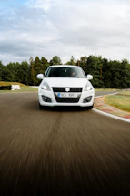 A white car is driving on a racetrack with trees and a cloudy sky in the background. The car has a license plate with a European design.