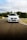 A white car is driving on a racetrack with trees and a cloudy sky in the background. The car has a license plate with a European design.