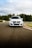 A white car is driving on a racetrack with trees and a cloudy sky in the background. The car has a license plate with a European design.