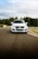 A white car is driving on a racetrack with trees and a cloudy sky in the background. The car has a license plate with a European design.