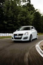A white car with a Suzuki emblem navigates a sharp curve on a road that is lined with blurred trees and a guardrail, indicating motion and speed.
