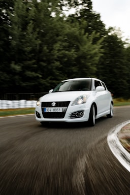 A white car with a Suzuki emblem navigates a sharp curve on a road that is lined with blurred trees and a guardrail, indicating motion and speed.