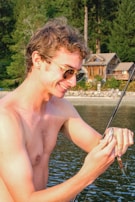 A shirtless man with sunglasses joyfully holds a fishing rod with a small fish caught on the line. In the background, there is a serene lakeside setting with lush green trees and a wooden cabin.
