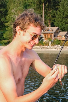 A shirtless man with sunglasses joyfully holds a fishing rod with a small fish caught on the line. In the background, there is a serene lakeside setting with lush green trees and a wooden cabin.