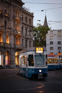 A blue and white tram marked with the number 13 and the destination Albisgütli moves along a curved track on a city street. The tram passes by a historic building with ornate architecture and warmly lit windows. Overhead, there are wires for the tram, and in the background, a church steeple rises above the buildings.