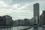 A rainy urban scene featuring a river bordered by railings on one side and a row of varied-height buildings on the other. A tall, modern skyscraper dominates the right side, while older apartment buildings line the left. Several vehicles, including a van, are driving on the wet street beside the river. The sky is overcast, with dense clouds reflecting the gloomy weather.