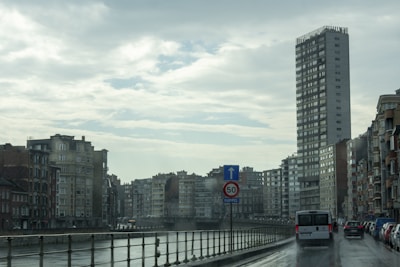 A rainy urban scene featuring a river bordered by railings on one side and a row of varied-height buildings on the other. A tall, modern skyscraper dominates the right side, while older apartment buildings line the left. Several vehicles, including a van, are driving on the wet street beside the river. The sky is overcast, with dense clouds reflecting the gloomy weather.
