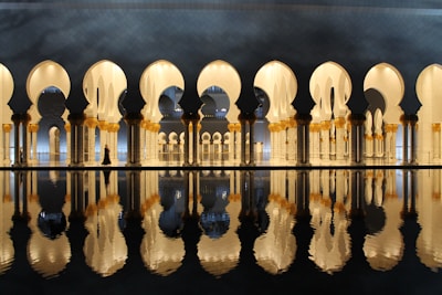 A series of ornate arches with intricate designs reflected in a calm body of water. The architectural elements feature gold and white colors, with the scene illuminated by soft lighting, creating a serene and majestic atmosphere.