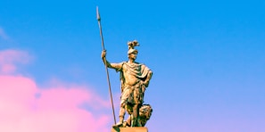 A bronze statue of a warrior stands holding a long spear in one hand and wearing a helmet adorned with a crest. The statue is set against a vibrant sky transitioning from blue to pink.