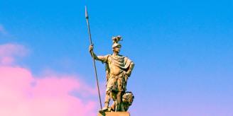 A bronze statue of a warrior stands holding a long spear in one hand and wearing a helmet adorned with a crest. The statue is set against a vibrant sky transitioning from blue to pink.