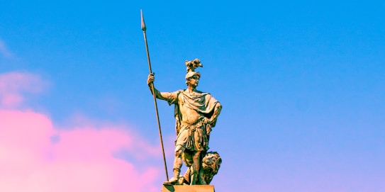 A bronze statue of a warrior stands holding a long spear in one hand and wearing a helmet adorned with a crest. The statue is set against a vibrant sky transitioning from blue to pink.