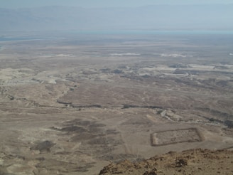 A vast desert landscape stretches out under a clear sky, with rugged terrain and a mix of light and dark sandy colors. The horizon features distant hills and a subtle blue hue of a water body. The scene includes a rectangular archaeological feature in the foreground, possibly a ruin or an ancient structure.