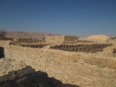 A vast archaeological site featuring ancient stone structures with rugged rock walls. The architecture is set against a backdrop of barren hills under a clear blue sky.