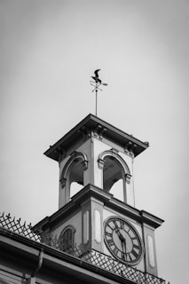 A clock tower with a traditional architectural design, featuring a large clock face with Roman numerals. The tower has arched openings and is topped with a small weather vane shaped like a bird. The black and white color scheme gives it a classic, timeless feel.