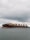 A large cargo ship with numerous shipping containers stacked on its deck, moving through a body of water under an overcast sky. The ship's hull is dark with a rusty red color near the waterline. The water appears somewhat choppy, indicative of light winds or a breeze.