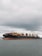 A large cargo ship with numerous shipping containers stacked on its deck, moving through a body of water under an overcast sky. The ship's hull is dark with a rusty red color near the waterline. The water appears somewhat choppy, indicative of light winds or a breeze.