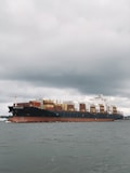 A large cargo ship with numerous shipping containers stacked on its deck, moving through a body of water under an overcast sky. The ship's hull is dark with a rusty red color near the waterline. The water appears somewhat choppy, indicative of light winds or a breeze.