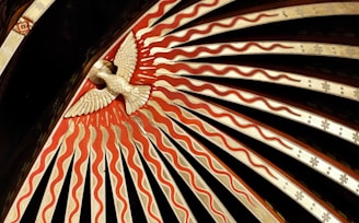 An ornate decorative design features a stylized bird in the center, surrounded by radiating patterns of red and beige waves. The bird appears to be carved or molded, positioned against a dark background with a series of intricately detailed rays emanating outward.