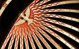 An ornate decorative design features a stylized bird in the center, surrounded by radiating patterns of red and beige waves. The bird appears to be carved or molded, positioned against a dark background with a series of intricately detailed rays emanating outward.