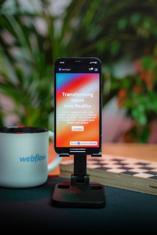 A smartphone is displayed on a black stand, showing an app with the text 'Transforming Ideas into Reality'. Behind the phone, there are blurred green plants. To the left, a white mug with 'webflow' written on it is placed. The scene is set against a dark surface, and there is a checkered board partially visible.