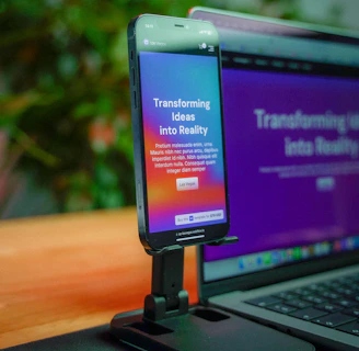 A smartphone is mounted on a stand, displaying a website with colorful graphics and the text 'Transforming Ideas into Reality'. Beside it, a laptop screen shows a blurred version of the same website. The background is soft and out of focus, with green foliage adding a hint of nature.