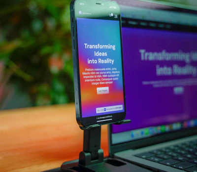 A smartphone is mounted on a stand, displaying a website with colorful graphics and the text 'Transforming Ideas into Reality'. Beside it, a laptop screen shows a blurred version of the same website. The background is soft and out of focus, with green foliage adding a hint of nature.