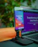 A smartphone is mounted on a stand, displaying a website with colorful graphics and the text 'Transforming Ideas into Reality'. Beside it, a laptop screen shows a blurred version of the same website. The background is soft and out of focus, with green foliage adding a hint of nature.