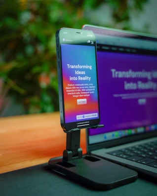 A smartphone is mounted on a stand, displaying a website with colorful graphics and the text 'Transforming Ideas into Reality'. Beside it, a laptop screen shows a blurred version of the same website. The background is soft and out of focus, with green foliage adding a hint of nature.