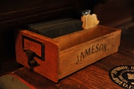 A wooden box with the name 'Jameson' printed on the side. The box contains dark-colored square coasters and some cream-colored napkins. It has a metal handle attached to the front.