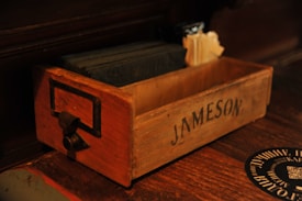 A wooden box with the name 'Jameson' printed on the side. The box contains dark-colored square coasters and some cream-colored napkins. It has a metal handle attached to the front.