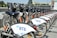 A row of rental bicycles are neatly lined up, showcasing a public bike-sharing system. The bikes have white fenders with the logo and design on them. They are parked in a designated area beside a concrete ledge, and the background features some greenery and city buildings.