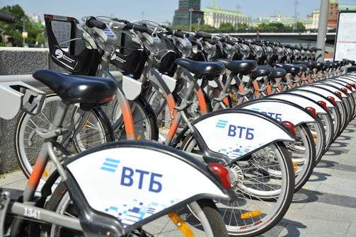 A row of rental bicycles are neatly lined up, showcasing a public bike-sharing system. The bikes have white fenders with the logo and design on them. They are parked in a designated area beside a concrete ledge, and the background features some greenery and city buildings.