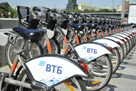 A row of rental bicycles are neatly lined up, showcasing a public bike-sharing system. The bikes have white fenders with the logo and design on them. They are parked in a designated area beside a concrete ledge, and the background features some greenery and city buildings.