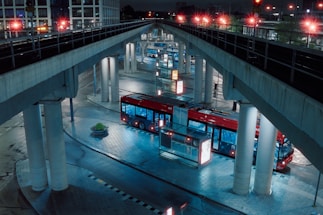 A red bus is parked in an urban transit station under several overpass bridges. The setting is at night, with artificial lights illuminating the scene, casting reflections on the wet, tiled pavement. The bus is surrounded by tall pillars and there is minimal activity around.