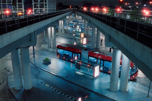 A red bus is parked in an urban transit station under several overpass bridges. The setting is at night, with artificial lights illuminating the scene, casting reflections on the wet, tiled pavement. The bus is surrounded by tall pillars and there is minimal activity around.