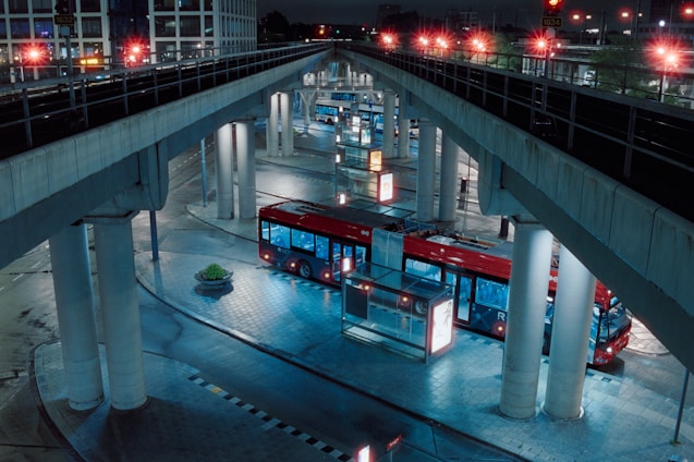 A red bus is parked in an urban transit station under several overpass bridges. The setting is at night, with artificial lights illuminating the scene, casting reflections on the wet, tiled pavement. The bus is surrounded by tall pillars and there is minimal activity around.