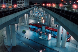 A red bus is parked in an urban transit station under several overpass bridges. The setting is at night, with artificial lights illuminating the scene, casting reflections on the wet, tiled pavement. The bus is surrounded by tall pillars and there is minimal activity around.
