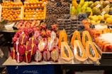 A vibrant display of exotic fruits in a market setting. Front and center are halved papayas, their seeds exposed, alongside vivid red and white dragon fruits with spoons placed inside their halves. Surrounding these are stacks of fresh oranges, dates, peaches, and other tropical fruits like mangoes and passion fruits. Each type is neatly organized with labels displaying their prices.